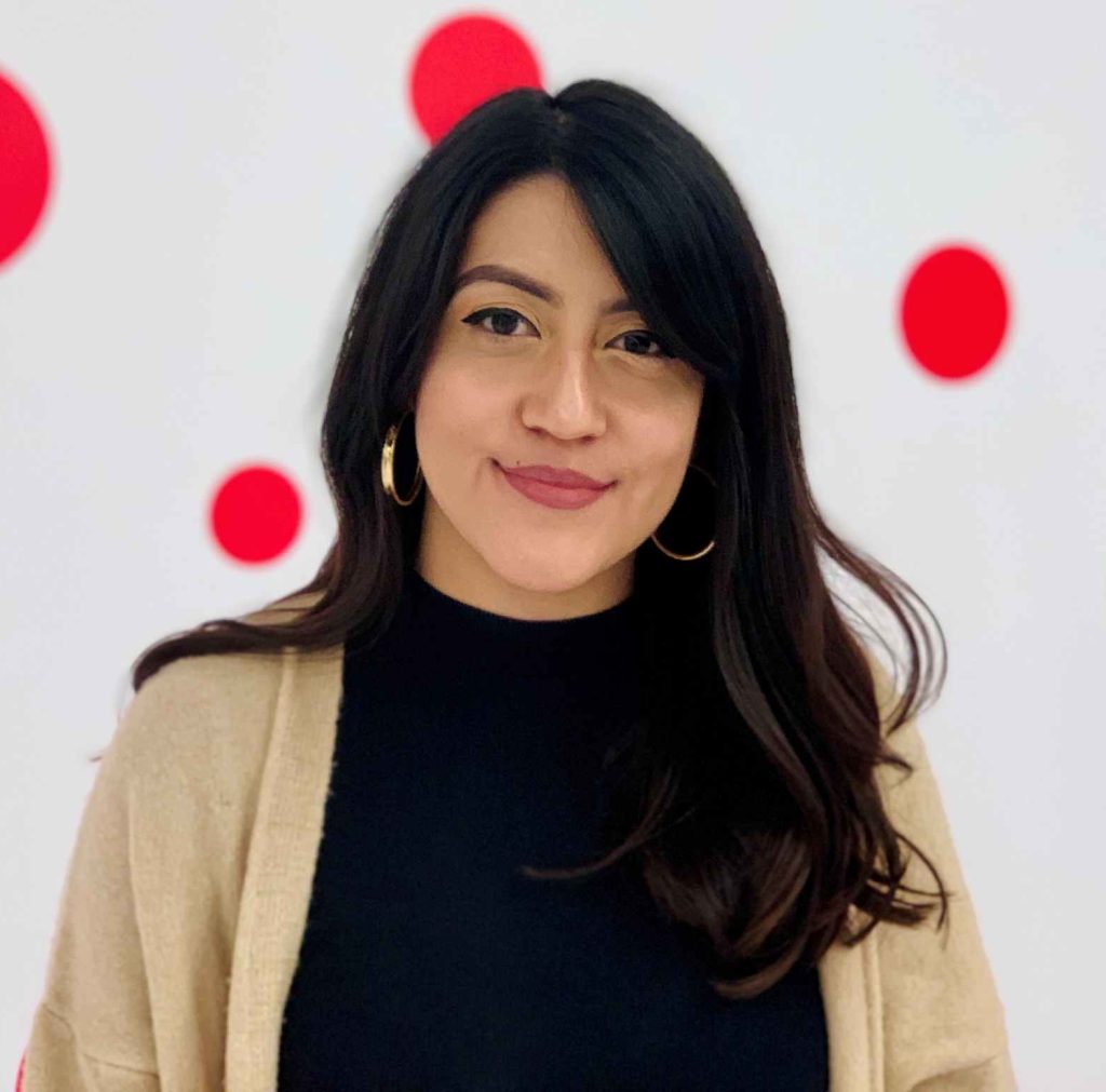 Portrait of a smiling woman with long dark hair, wearing a black top and beige cardigan, against a white backdrop with red circles.